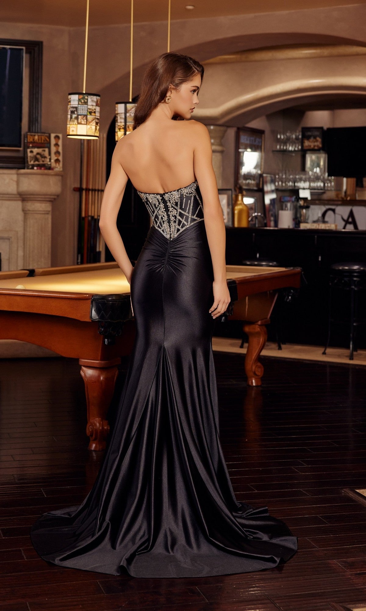 A woman faces away from the camera in an upscale indoor setting by a pool table, wearing the Nox Anabel E1901 Long Prom Dress featuring a strapless satin skirt and crystal beaded embellishments.